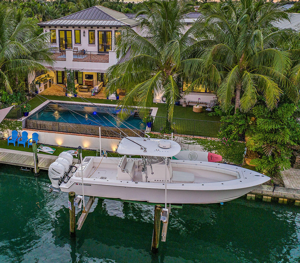 Boat docked by a luxurious house with a pool, surrounded by palm trees. Blue chairs visible; serene tropical setting at dusk.