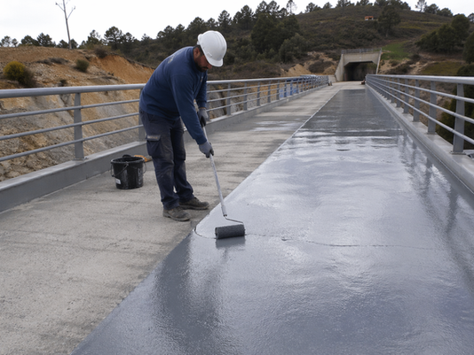 Foto de la aplicación de membrana de impermeabilización sobre el tablero de una pasarela peatonal de hormigón — operario aplicando el sistema con rodillo, superficie gris de hormigón visible a los lados. Ambiente de obra civil exterior. Transmite escala y contexto técnico real.