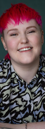 Headshot of Lozza, a white non-binary person with a bright pink mullet wearing a funky shirt that is green, blue and black. They're smiling like they're mid-laughter