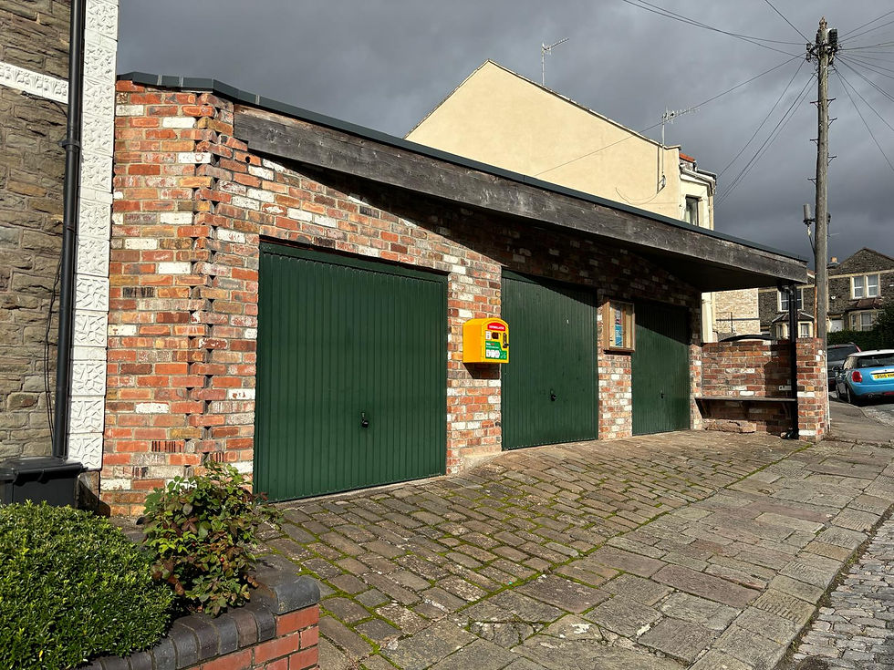 Bright yellow defibrillator mounted on a brick wall between garages on Islington Road, Southville, providing 24-hour access to emergency life-saving equipment