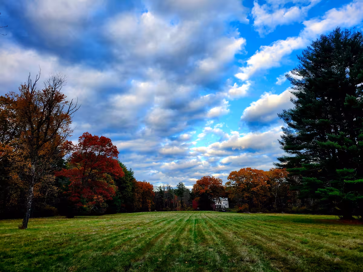 Green field mowed in rows with fall foliage surrounding and partly cloudy sky.