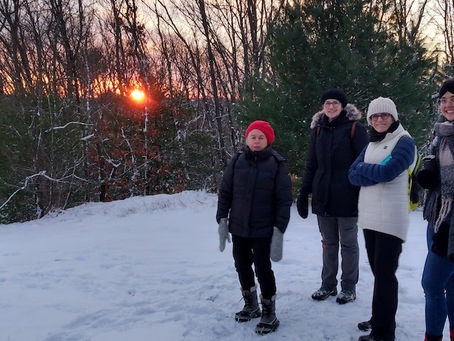Hikers atop snowy Cat Rock on the a past winter solstice hike, with the sun just rising on the horizon behind them, screened by trees.