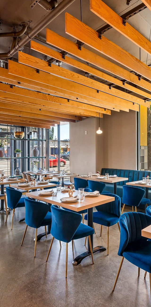 Modern restaurant interior with natural wood ceiling beams, floor-to-ceiling windows, and blue velvet dining chairs.