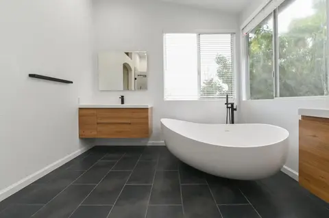Minimalist bathroom with white freestanding soaking tub, wood floating vanity, black floor tiles, and large windows – Green Bay Remodeling