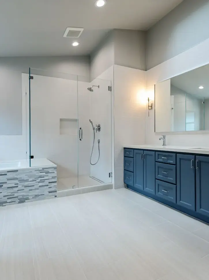 Spacious bathroom with navy blue double vanity, frameless glass shower, soaking tub with gray mosaic tile, and light porcelain floors – Green Bay Remodeling