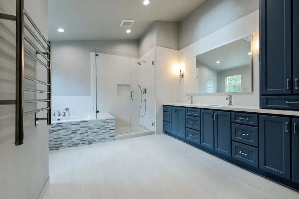 Spacious bathroom with navy blue double vanity, frameless glass shower, soaking tub with gray mosaic tile, and light porcelain floors – Green Bay Remodeling
