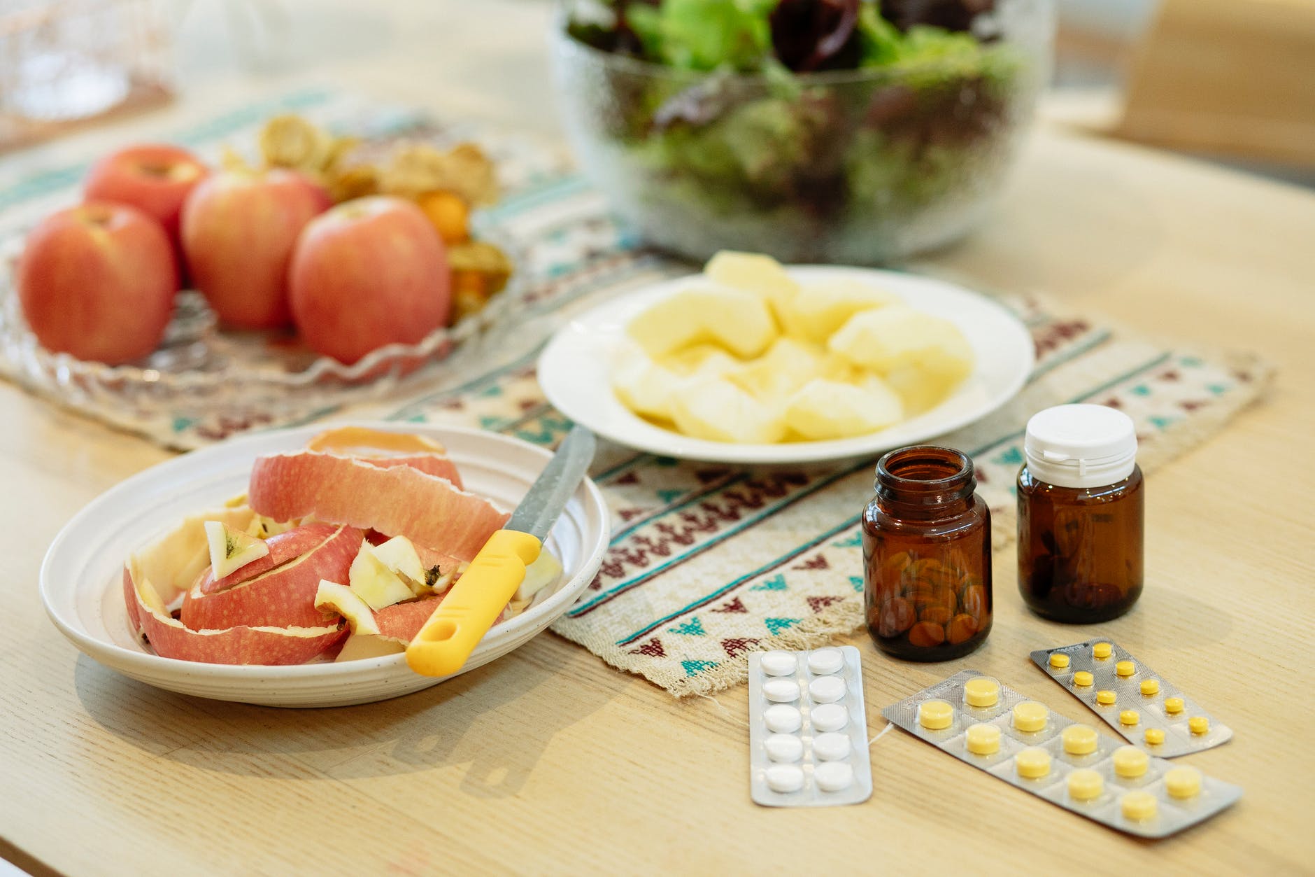 Sliced apples on a plate with a knife and whole apples nearby. On a tablecloth, jars and pill packs are arranged, salad in the background.