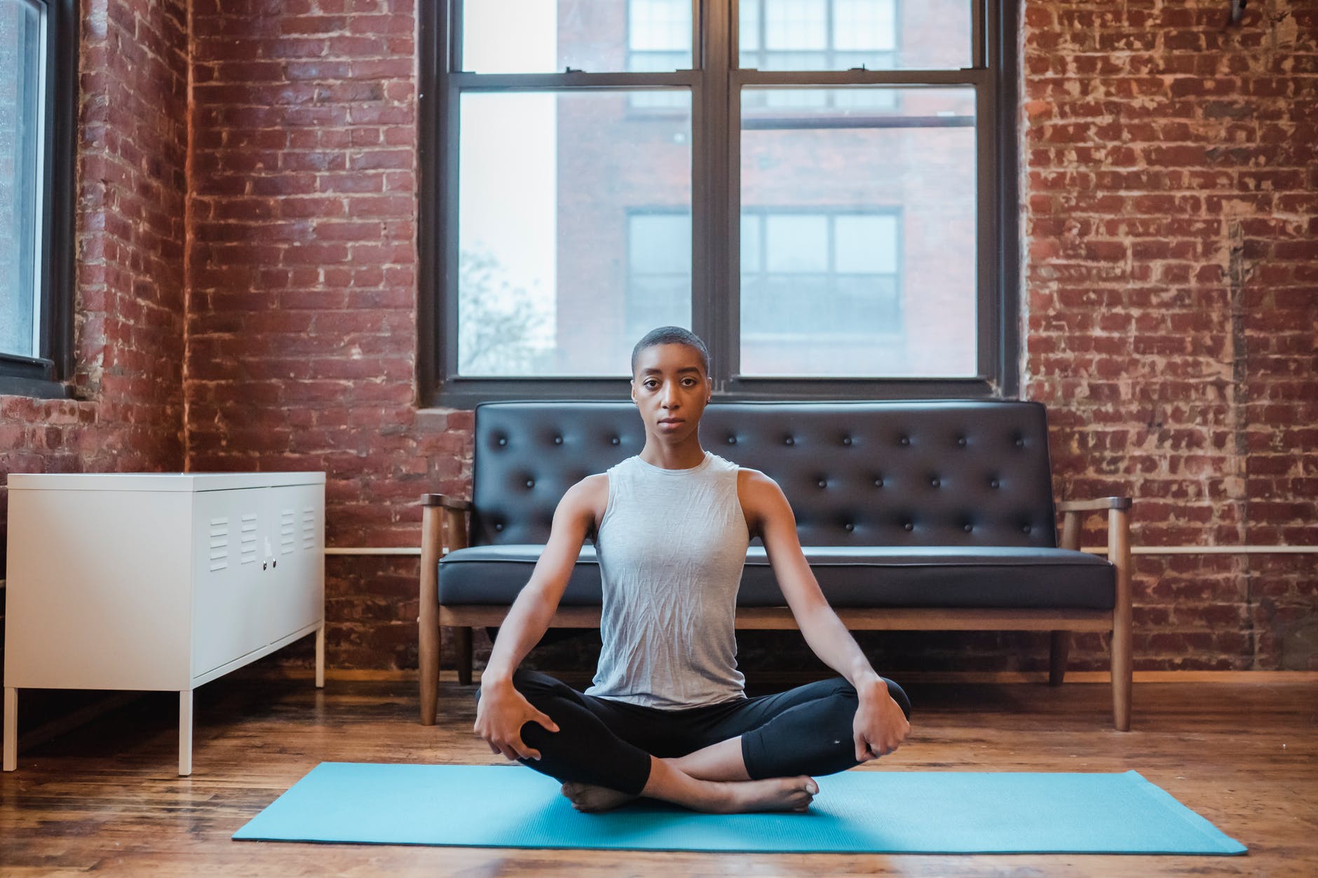 Person in gray tank top meditates on a blue yoga mat in a brick-walled room with a black sofa, wooden floor, and large windows. Calm mood.