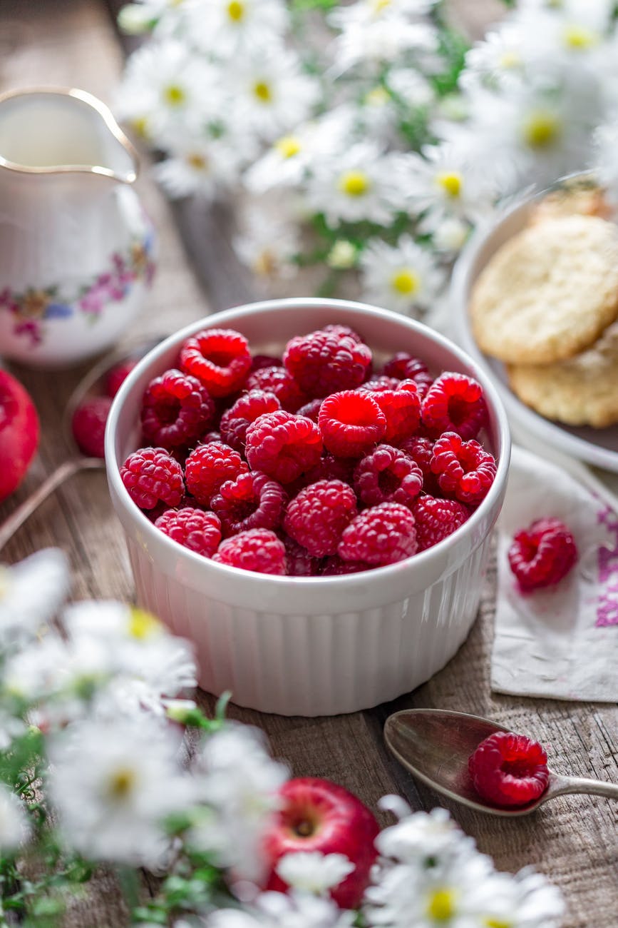 Bowl of fresh red raspberries on wooden table with daisies, a spoon, a pitcher, and cookies. Bright, rustic, and inviting scene.