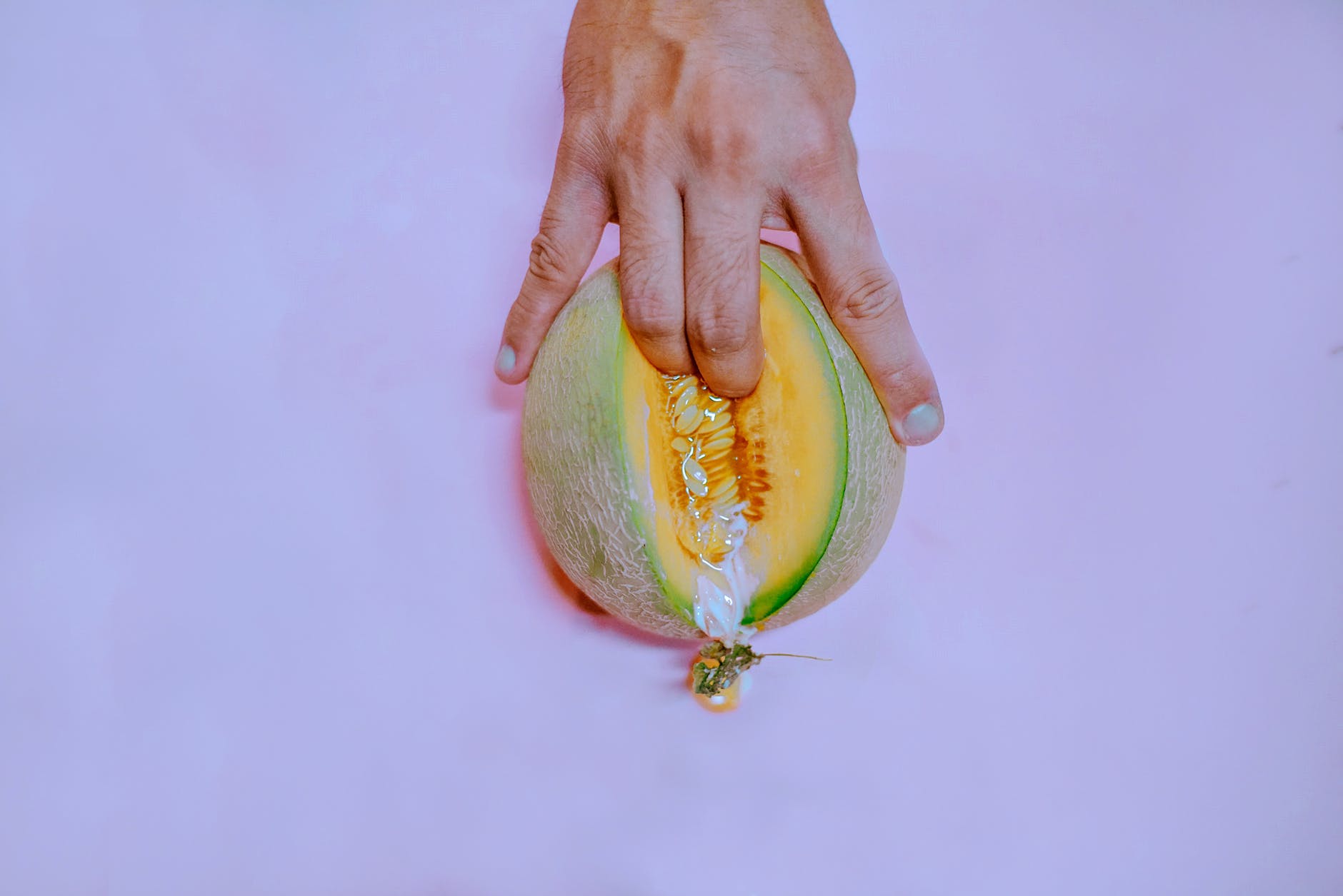 Hand pressing open a melon on a pink background, revealing orange flesh and seeds. Vibrant colors and playful mood.