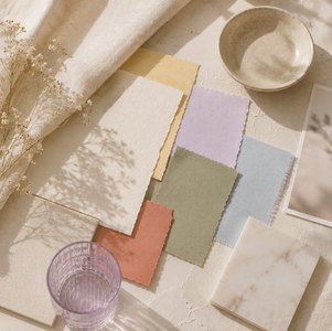 Soft pastel fabric swatches on cream surface, with a delicate plant, marble tile, bowl, and glass, creating a serene and natural mood.