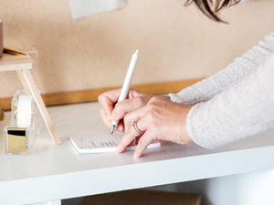 Hands writing on a notepad with a pen, beside a calculator, tape dispenser, and cup in a neutral-toned office space.