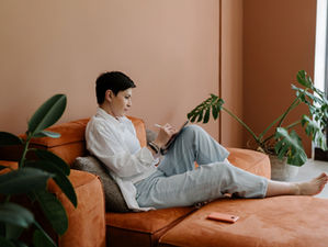 A person in a white shirt uses a laptop on an orange couch in a room with peach walls and large green plants, creating a relaxed mood.