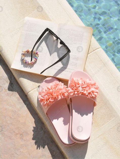 Pink floral slides, sunglasses, and an open book on a poolside lounger beside blue water in bright sun.