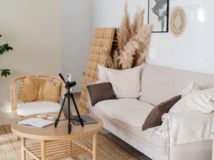 Cozy living room with a beige sofa, brown pillows, and a small table holding a tripod, phone, laptop, and notebook. Plants and neutral decor.