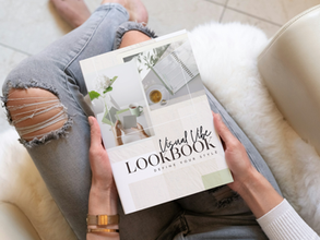 Hands hold "Visual Vibe Lookbook" on lap in ripped jeans. White and beige setting with a cozy mood. Book cover shows design images.