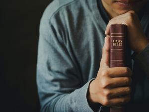 Person in gray sweater holding a closed red Bible with "HOLY BIBLE" text, in a dim setting, conveying contemplation or prayer.