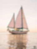 Pink checkered-sail sailboat with an American flag glides on calm water at sunset, reflecting softly.