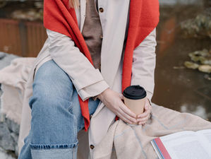 Person in jeans and a red scarf sits by a pond, holding a coffee cup. A book lies open on their lap. Calm, autumn setting.