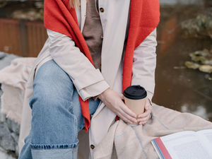 Person in jeans and a red scarf sits by a pond, holding a coffee cup. A book lies open on their lap. Calm, autumn setting.