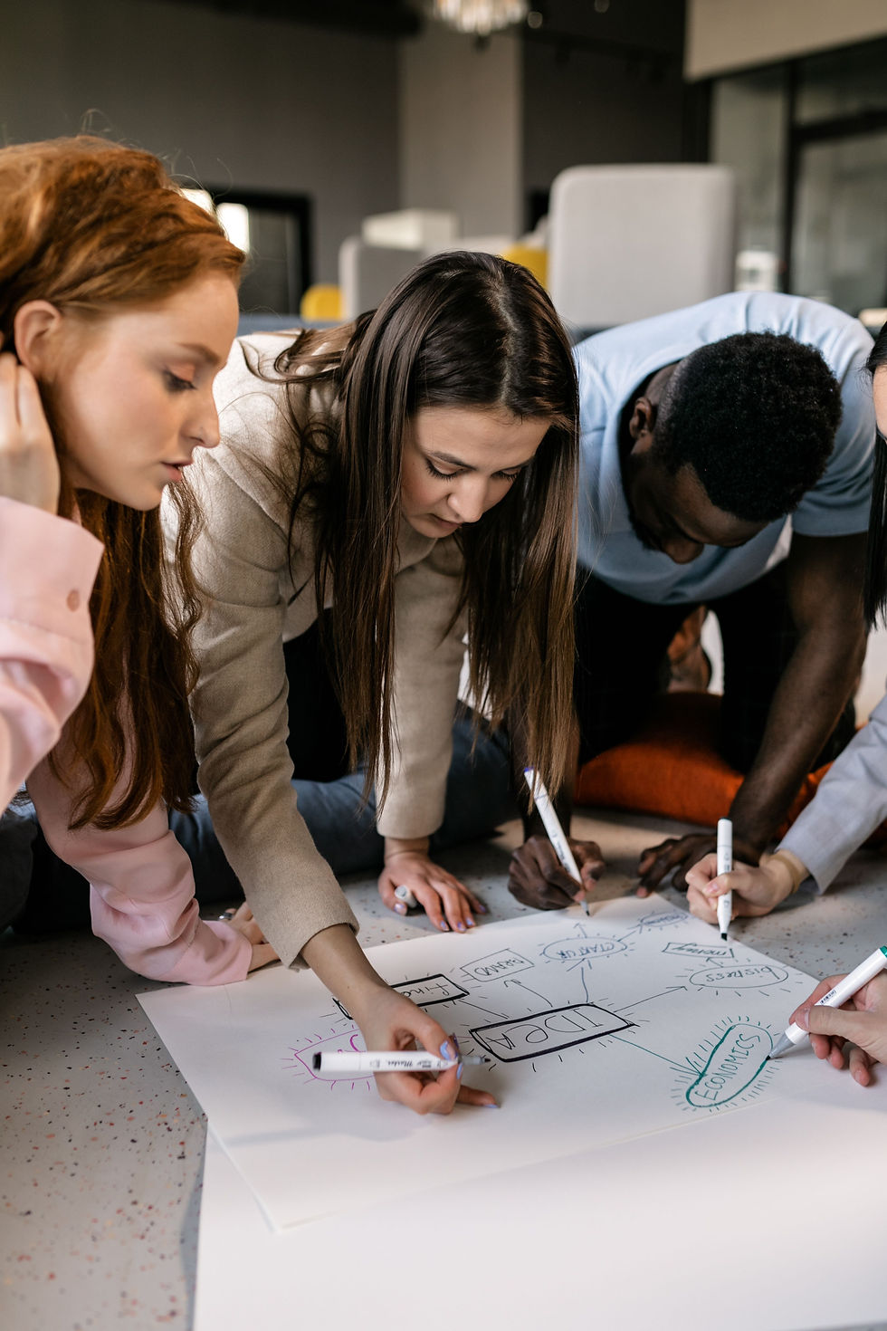 Diverse coworkers lean over a floor poster brainstorming and writing ideas like IDEA and ECONOMICS in a bright office.