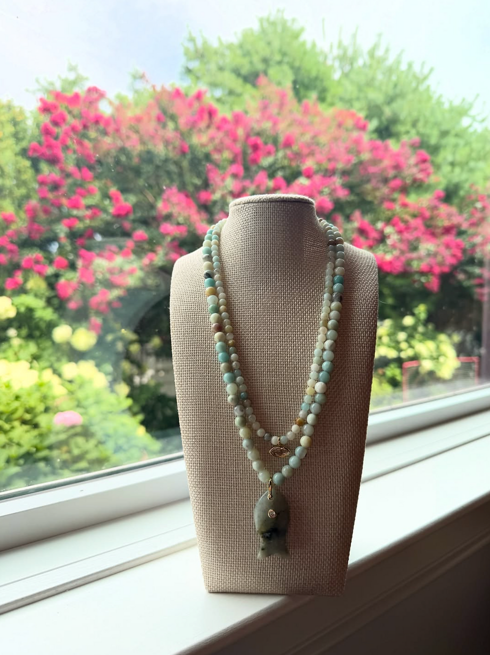 Beaded necklace with fish pendant on a linen bust by a window, with pink flowering trees blurred outside.
