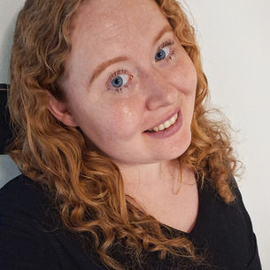 Smiling red-haired woman in a black top poses against a plain white wall, looking at the camera.