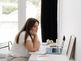 Thoughtful woman in a bright home office sits at a desk with a laptop, notebook, phone, and mug, looking down.