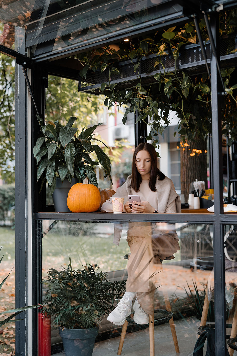 Woman sits in a plant-filled café, looking at her phone beside a pumpkin and paper cup, with a calm, autumnal mood.