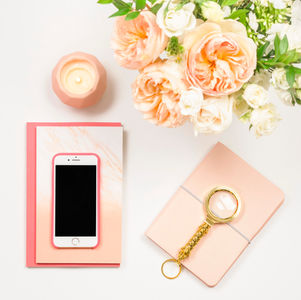 Peach and white flowers, candle, pink phone on a notebook, magnifying glass on a beige journal on white desk; elegant and calm mood.