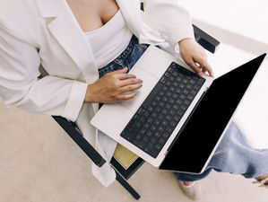 woman in white blazer using a laptop on their lap, seated in a chair. The setting is bright, with neutral tones and casual attire.