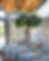Elegant dining table set with blue napkins, gold cutlery, and candles. Red roses in vases. Large windows and tree in the background. Warm lighting.