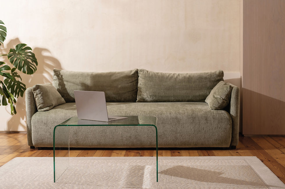 Laptop on a glass table in front of a green sofa. Wooden floor, beige wall, and a large plant add to the cozy, minimalistic setting.
