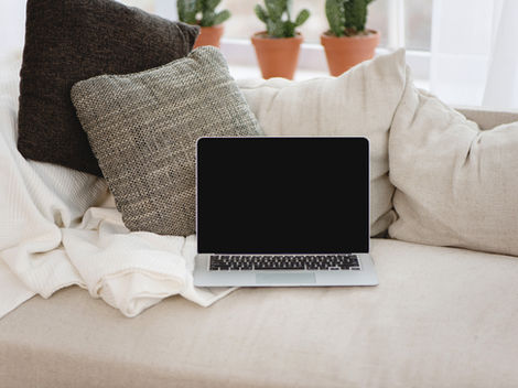 Laptop on a beige couch with textured pillows, surrounded by blankets and small potted cacti on a windowsill. Cozy home setting.