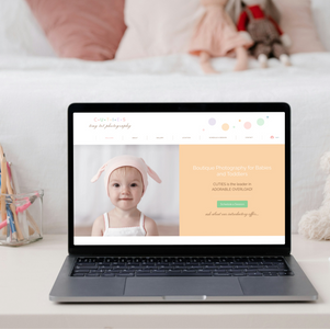Laptop displaying photography website with baby image. White, cozy bedroom setting with teddy bear, pencils in jar, and books nearby.
