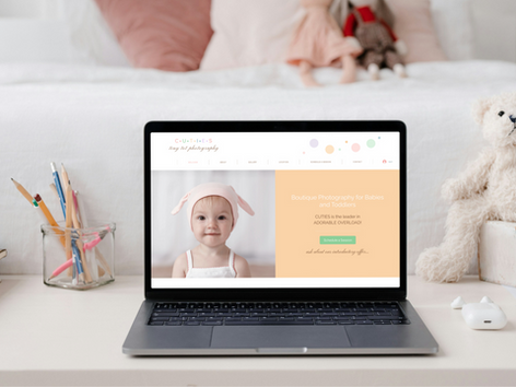 Laptop displaying photography website with baby image. White, cozy bedroom setting with teddy bear, pencils in jar, and books nearby.