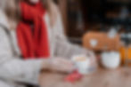 Woman in beige coat and red scarf holds a cup at a wooden table. A red leaf, teapot, and sugar bowl are nearby, creating a cozy mood.