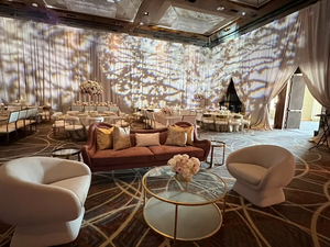 Elegant event room with plush seating, round tables, floral centerpieces, and soft lighting. Curtains and carpet feature geometric patterns.