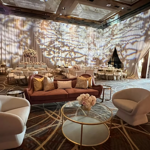 Elegant event room with plush seating, round tables, floral centerpieces, and soft lighting. Curtains and carpet feature geometric patterns.