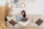 Woman in a light blue shirt sits cross-legged on a beige couch using a laptop. Minimalist room with plants, artwork, and neutral tones.