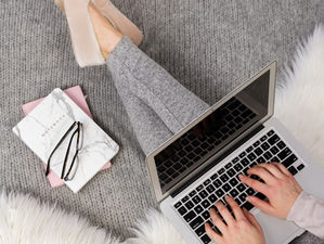 Person typing on a laptop, wearing gray pants and pink slippers. Notebooks and glasses beside them on a gray carpet with white fur.
