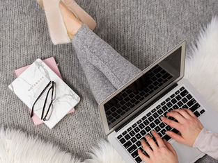 Person typing on a laptop, wearing gray pants and pink slippers. Notebooks and glasses beside them on a gray carpet with white fur.