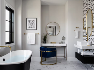 Modern bathroom with a black tub, blue chair, and geometric wall pattern. Gold fixtures, round mirror, and marble floor add elegance.