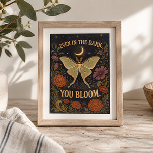 Framed botanical print with a luna moth and crescent moon reading EVEN IN THE DARK, YOU BLOOM, on a rustic table with vase and bowl