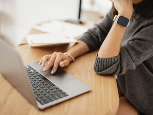 Person in a gray sweater using a laptop on a wooden desk, wearing a smartwatch. Papers and a phone in the background convey focus.