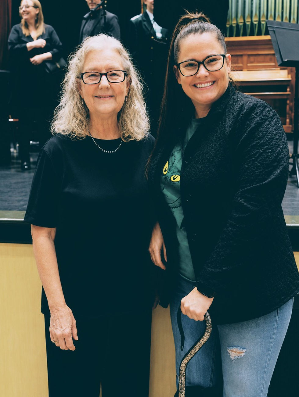 Smiling older and younger women with glasses stand indoors, the younger holding a snake; stage and organ behind them.