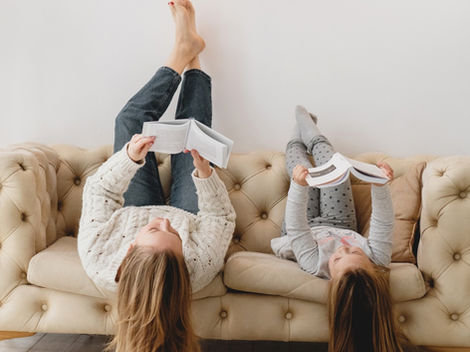 Two girls lie upside down on a beige sofa, reading books in a bright modern living room with wooden pendant lights and a plant.