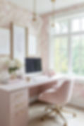 Soft pink home office with a computer, rose chair, floral wallpaper, gold pendant lights, and a bright window with trees outside.