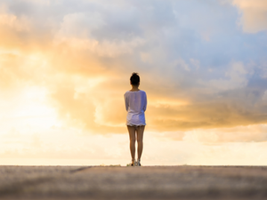 Person standing alone facing a bright sunset, wearing a white shirt and shorts, with a vast sky and clouds in the background. Serene mood.