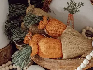 Fabric carrots with green tops in a wooden bowl, surrounded by wooden beads and decor with carrot image. Rustic and cozy setting.